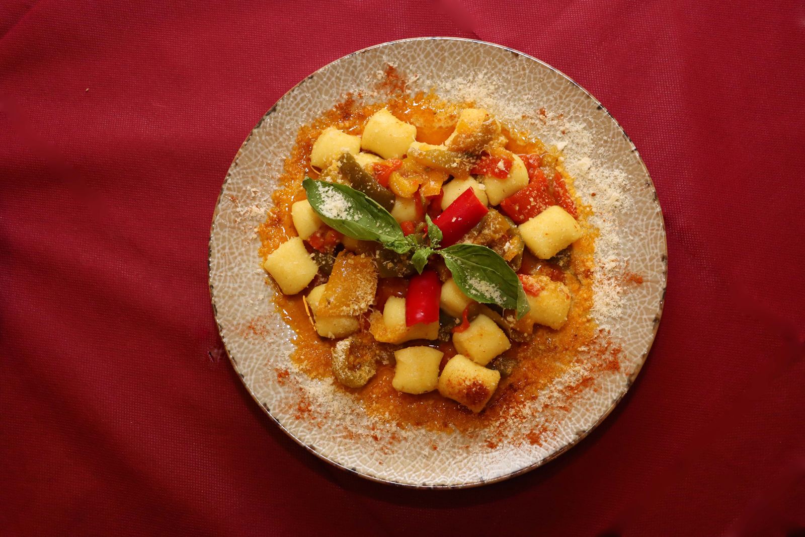 Piatto di gnocchi ai peperoni rossi e gialli con foglie di basilico e formaggio grattugiato, visto dall'alto sulla tovaglia bordeaux