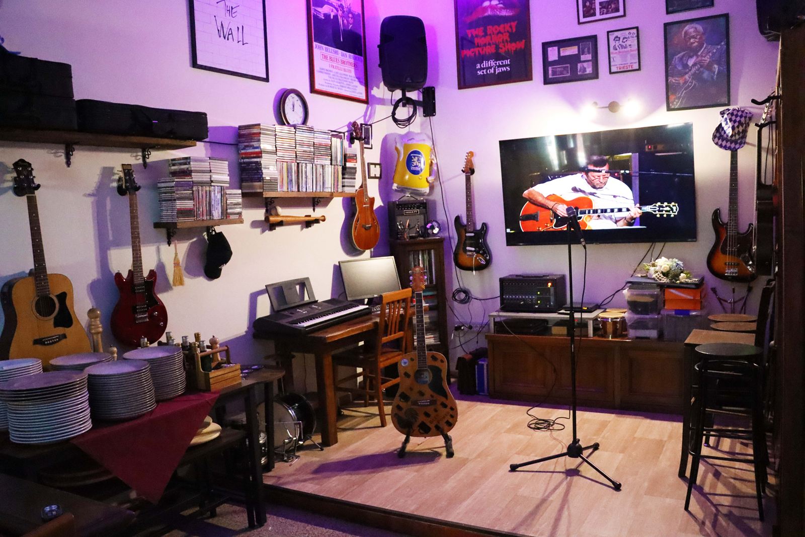 L'angolo musicale del buffet con luci viola: chitarre appese alle pareti, una tastiera, un amplificatore, un'asta del microfono e un televisore con un concerto
